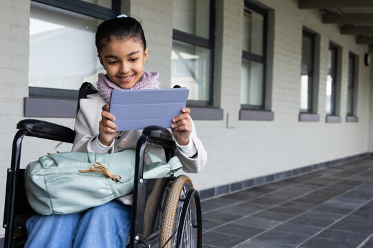 African-descent child girl sitting in wheelchair using blue tablet pack on lap corridor, copy space