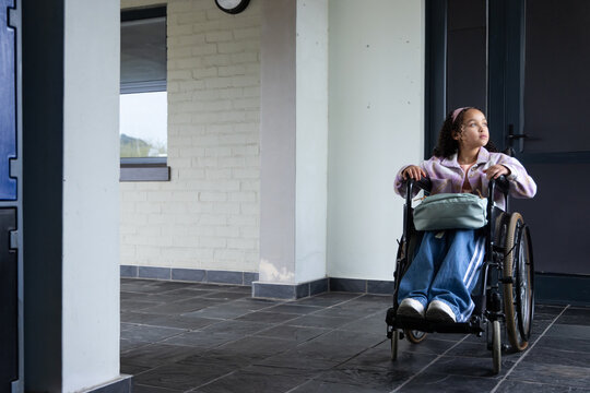 Youth African female sitting manual wheelchair in foyer wearing pink jacket, bag on lap, copy space