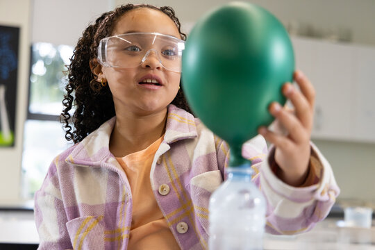 African American teen girl wearing plaid jacket and goggles holding green balloon on bottle in lab