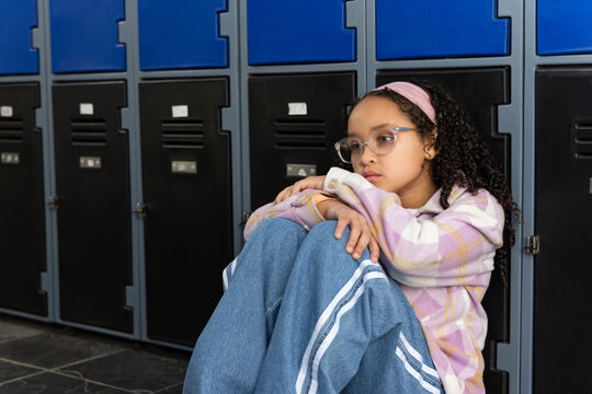 Teenage female sitting in school hallway hugging knees wearing pink headband by lockers, copy space