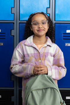 African American girl child standing at blue lockers wearing pink headband holding green backpack