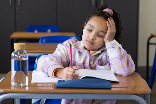 African American child sitting at school desk with pink scrunchie, writing notebook with red pencil