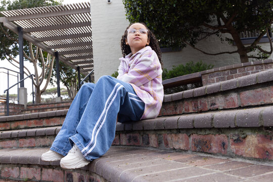Teenage African female sitting on brick steps in courtyard wearing pastel jacket and glasses