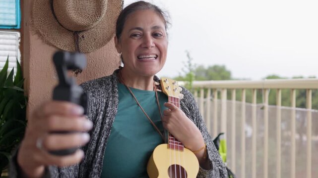 middle eastern senior woman holding a ukulele while filming a video blog with a vlogging camera on her home balcony, smiling and talking to her audience with genuine happiness and engagement