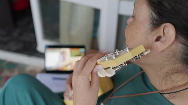 Middle eastern woman learning to play the ukulele through an online video lesson on her laptop. Practicing chords and strumming in her living room.hobby and developing a new musical skill