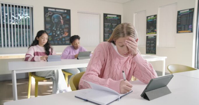 Diverse female pupils in class task urging front blond writing spiral notebook, pen beside tablet