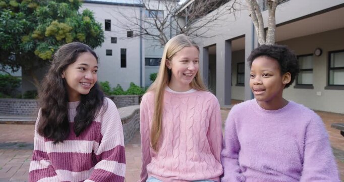 Diverse teen girl friends in sweaters center speaking smiling leaning in, posing on courtyard bench