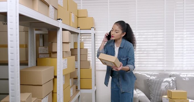 Asian woman entrepreneur holds parcel box and speaks on smartphone, explains order availability to customer, coordinates e-commerce packaging workflow and delivery schedule