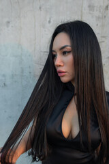 Profile portrait of a gorgeous Asian woman with long black hair, wearing a modern black keyhole dress, looking away thoughtfully against a textured concrete wall during daylight. Pretty sexy lady © alones