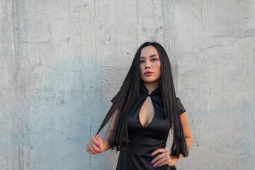 Fototapeta premium Confident Asian woman with long black hair, wearing a modern black cheongsam dress, looking at camera against a textured concrete wall, hand on hip. Pretty glamour urban elegant girl