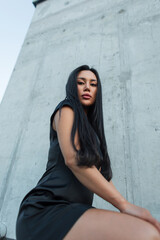 Fototapeta premium Low angle portrait of a confident Asian woman with long black hair looking at the camera, wearing an elegant black dress against a tall concrete wall. urban fashion photography. Pretty hot lady