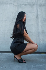 Side profile portrait of a stylish Asian woman with long black hair crouching in a short black silk dress and high heels against a concrete wall. Chic urban fashion look. © alones