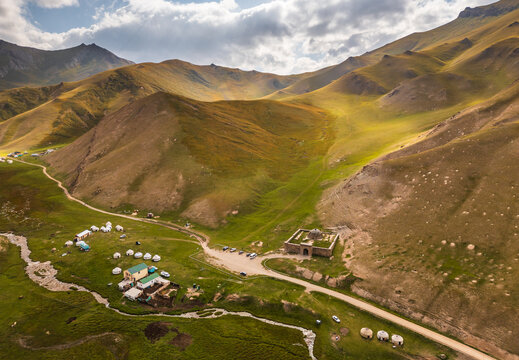 Aerial view of Tash Rabat Caravan Palace in a lush green valley surrounded by mountains, with traditional yurts and a winding stream in At-Bashi District, Naryn Region, Kyrgyzstan.