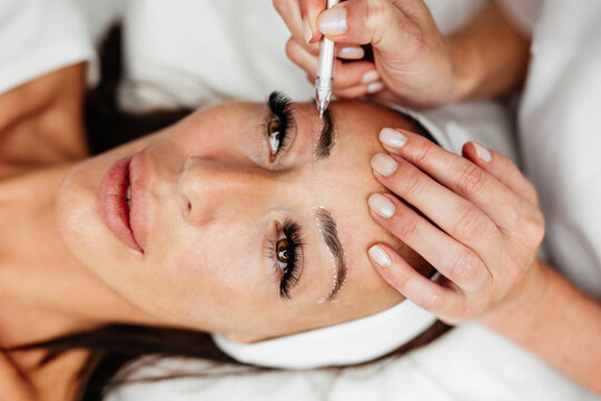 A professional beautician in a beauty salon marks symmetry lines on a woman's forehead before permanent makeup treatment.