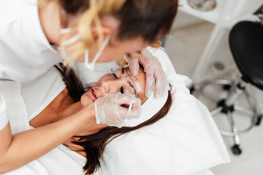 A professional beautician in a beauty salon marks symmetry lines on a woman's forehead before permanent makeup treatment.