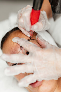 Close-up shot of a specialist applying semi-permanent makeup and pigment to a client's brow line using specialized equipment and sterile gloves during a beauty treatment.