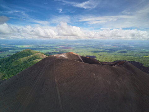 Aerial view of Cerro Negro volcano with its dark volcanic slopes, surrounded by lush green forests and rolling hills under a blue sky with white clouds in Larreynaga, Leon, Nicaragua.
