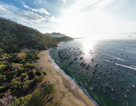 Aerial view of Papuma Beach with numerous traditional fishing boats anchored in the water along the sandy coastline, surrounded by lush green hills in Jember, East Java, Indonesia.