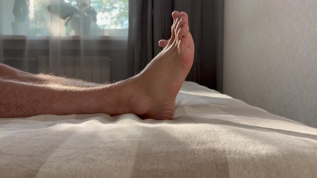 a man's hairy legs and bare feet resting on a textured bedspread in a sunny bedroom. Relaxation, leisure, and waking up on a lazy morning.