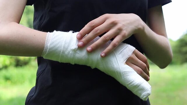 Injured person wraps a white medical bandage around their wrist for support while standing outside in a park on a bright day to help with healing and protect the skin