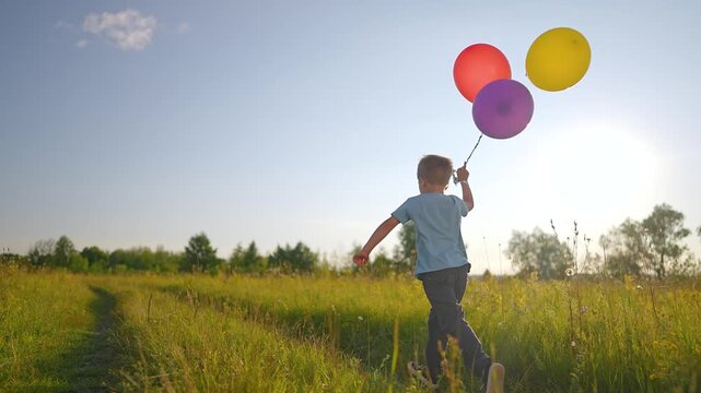 Boy walks through meadow holding colorful balloons. Child carries balloon bundle while walking in field. Kid walks at sunset. Boy holds balloons outdoor. Child walks with colorful balloon.
