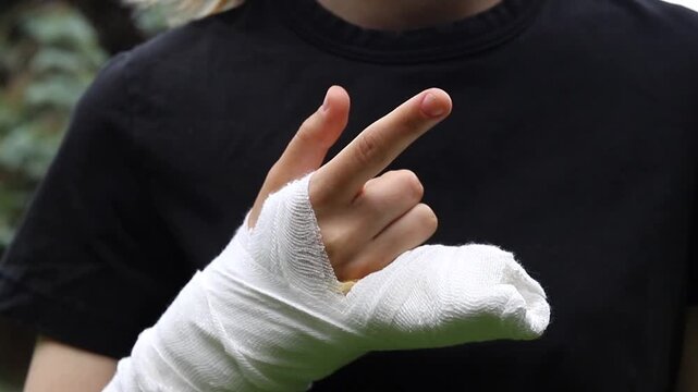 Broken hand wrapped in thick white gauze bandage shown outdoors in daylight as person makes a peace hand gesture while wearing black shirt