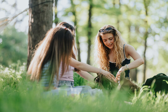 High school students sit on grass sharing backpacks and chatting outdoors. Friends enjoy a relaxed picnic-like moment in a sunlit park among trees.
