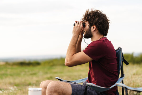 Man Sitting on a Camping Chair Using Binoculars to Observe Nature in a Grassy Field Near Hills
