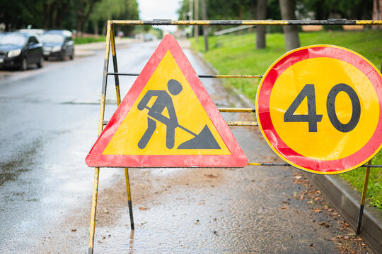 A construction zone is set up on a wet road with warning signs indicating ongoing work. The triangular sign shows a figure with a shovel, while the circular sign displays a speed limit of 40
