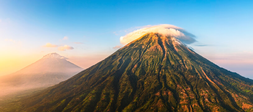 Aerial view of Mount Sindoro with a lenticular cloud cap over its peak, lush green volcanic slopes, and golden hour light in Kejajar, Central Java, Indonesia.