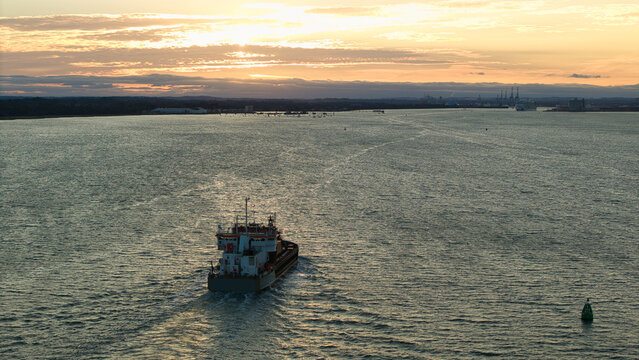 Aerial view of a cargo ship navigating the River Test at sunset with calm waters and a golden sky in Southampton, England, United Kingdom.