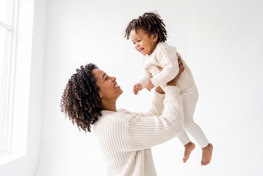 Happy Black Mother Lifting Her Laughing Toddler Daughter Up in the Air in a Bright Minimalist Room