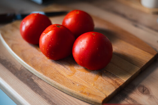 Four bright red tomatoes are placed on a wooden cutting board in a kitchen. Water droplets glisten on the tomatoes, showing their freshness and readiness for cooking