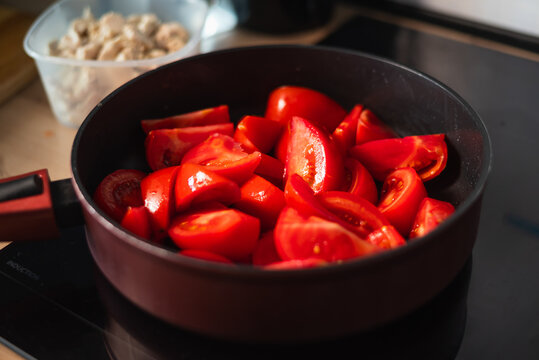Chopped fresh tomatoes are placed in a black pan on a stovetop in a kitchen. The scene captures the process of preparing ingredients for a meal
