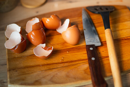 Brown eggshells are scattered on a wooden cutting board next to a knife and a spatula.