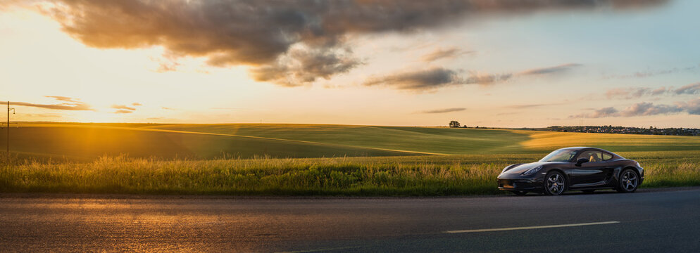panoramic view of Porsche Cayman 718 standing on the side of the road next to the hills of a field in the evening rays