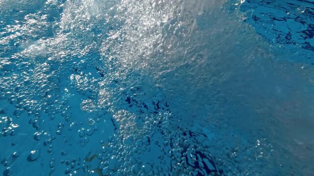 Detailed overhead shot of bubbles rising and spreading across the surface of clear swimming pool water near a circulation return outlet.