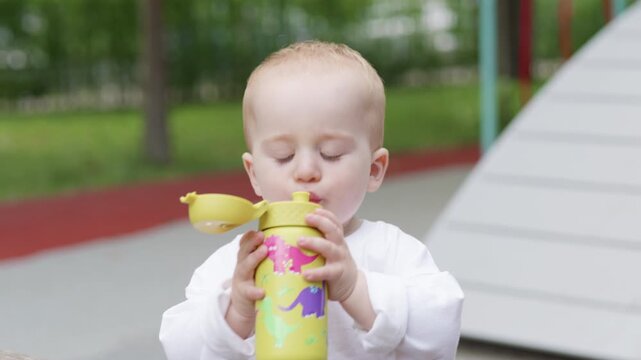 Caucasian toddler in white shirt plays with yellow sippy cup, lifting it to mouth and examining it while sitting at a playground with colorful background elements