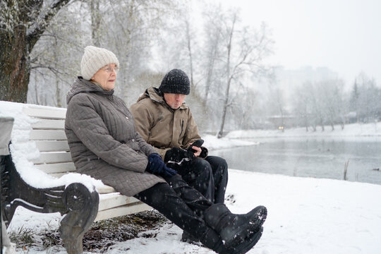 Senior couple sitting on bench looking at phone together in winter park. Elderly pair sharing modern moment on cold snowy day. Connected partners enjoying outdoor rest beside frozen lake