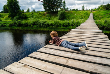 Obraz premium A focused young boy lies on a wooden bridge over a river, patiently fishing while relaxing in nature. This peaceful scene captures a child enjoying a rural retreat and observing the water.
