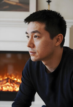 Thoughtful young asian man looking away in a cozy home setting with a fireplace in the background, a warm fireplace with glowing flames provides a comforting background