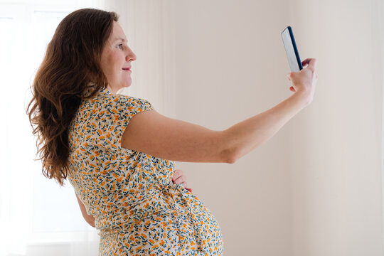 Pregnant woman holds her smartphone for selfie or video call, sharing happy moment indoors. Mother talking excitedly shows joyful, cheerful excitement and glowing maternal happiness at home
