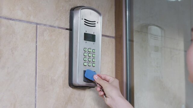 Hand holding blue access card approaches electronic keypad for entry, illuminated buttons visible, mounted on wall in modern indoor setting with tiled surface