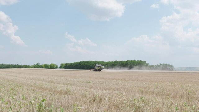 Agricultural machinery harvesting wheat in a vast field under a clear blue sky with scattered clouds, showcasing the continuous action of crop collection and dust cloud formation