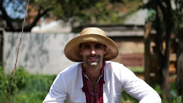 Portrait of ukrainian man in straw hat against background of summer garden. Young smiling guy wearing an embroidered shirt looking into camera outdoor. Concept of national identity and patriotism