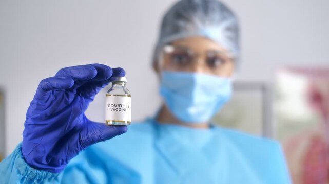 Medical staff wearing personal protective equipment kit showing COVID-19 vaccine. Female doctor in PPE kit holding a new effective vaccine against Coronavirus - flu shot vaccination drive