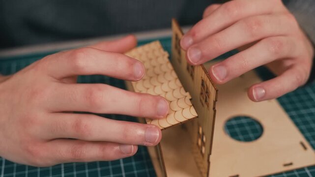 Attaching porch canopy to miniature wooden house model assembly. DIY architectural crafting featuring precise component fitting and handcrafted construction process. Shaping first home dream through
