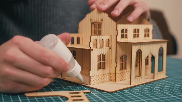 Crafting wooden house model with hands applying glue, installing final wall panel for DIY miniature architecture. New housing concept, creative home construction hobby, handmade real estate prototype