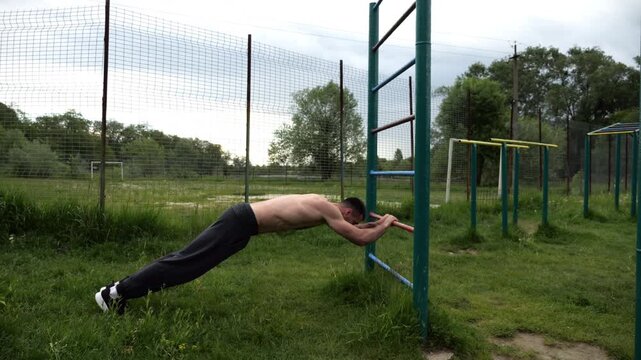 Young muscular athlete doing exercise on triceps at sport equipment outdoor. Male shirtless athlete working out at sport ground. Strong sporty man training at nature. Concept of healthy lifestyle