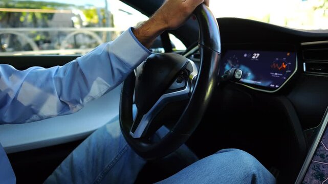 Male hand of businessman holds steering wheel while driving electrical vehicle at city. Businessperson operating electric car on urban road. Young man commuting to work at sunny day. Close up
